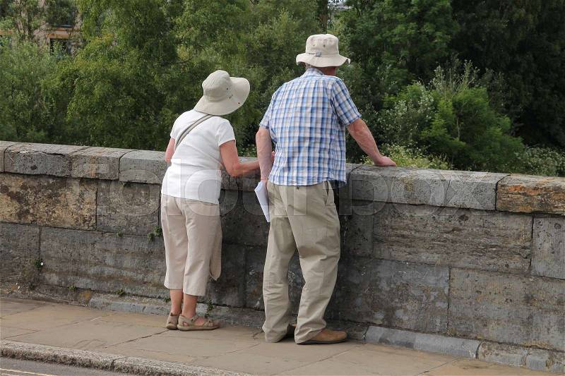 Retired couple, man and wife with hats, look over the wall from the bridge in the summer in England, stock photo
