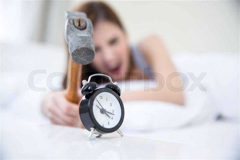 Woman not wanting to get up, taking a hammer to her alarm clock. Focus on clock, stock photo