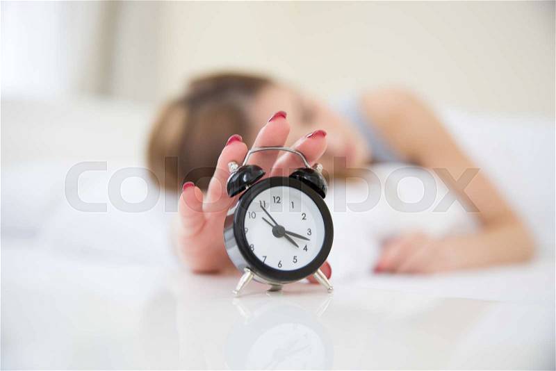 Sleepy woman in bed switching off alarm clock. Focus on clock, stock photo