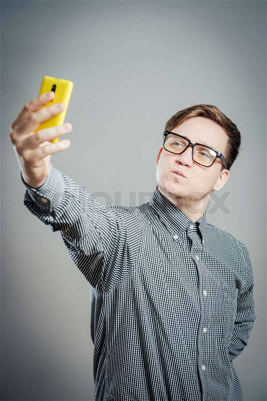 Handsome young man in shirt holding camera and making selfie and smiling while standing against grey background, stock photo