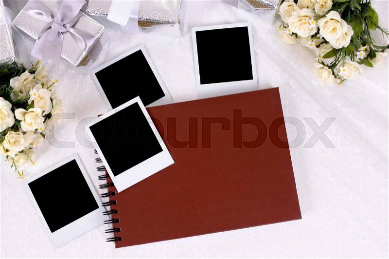 Red photo album with blank instant photo prints laid on bridal lace with several silver wedding gifts and white rose bouquets. Space for copy, stock photo