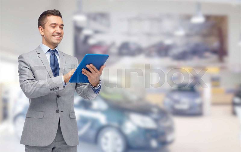 Business, people, car sale and technology concept - happy smiling businessman in suit holding tablet pc computer over auto show or salon background, stock photo