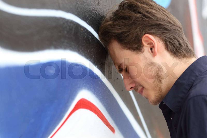 Depressed sad teenager boy lamenting with his head against a wall outdoors, stock photo