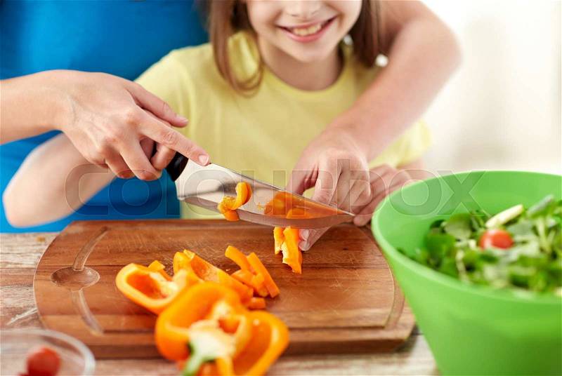 Food, family, cooking and people concept - close up of happy girl and mother making salad for dinner and chopping paprika on cutting board with knife in kitchen, stock photo