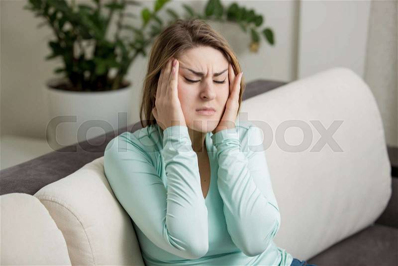 Young woman sitting on sofa and suffering from head ache, stock photo