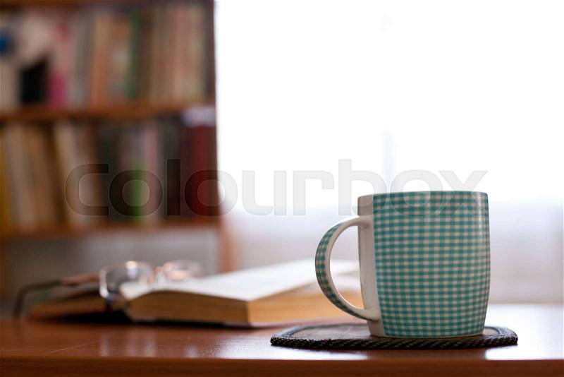 Cup with book on a table and shelf with books next to window on background, stock photo