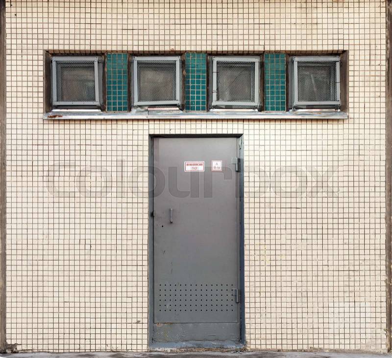 Industrial building wall texture with small square windows and metal door. Russian text on the label means flammable gas, stock photo