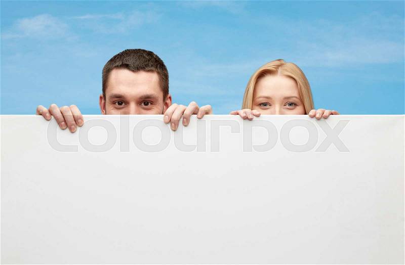People, advertisement and information concept - happy couple hiding behind big white blank board over blue sky background, stock photo