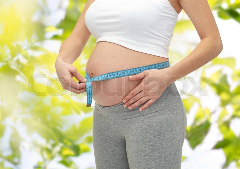 Pregnancy, motherhood, control, people and expectation concept - close up of happy pregnant woman measuring her bare tummy over green tree leavers background, stock photo