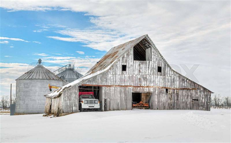 A snowy winter landscape surrounds a large, weathered white barn and farm equipment, stock photo