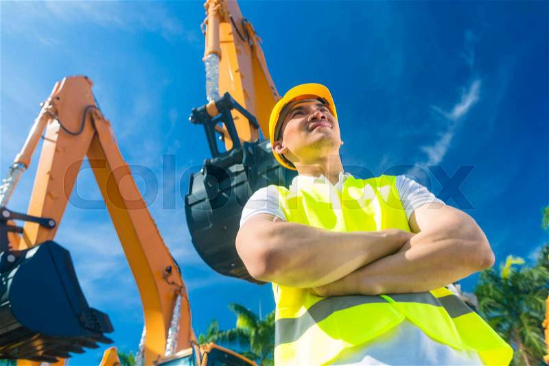 Asian worker standing in front of construction machinery of building site or rental company, stock photo