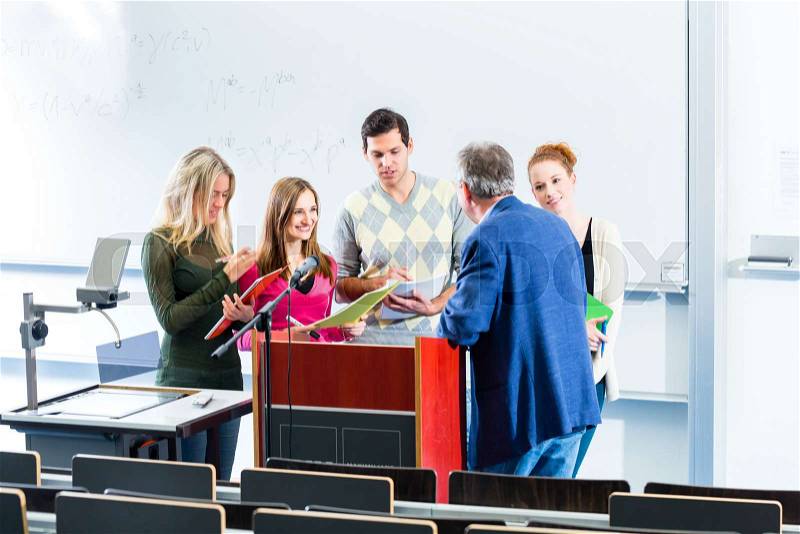 College students making lesson notes while asking professor in university auditorium, stock photo