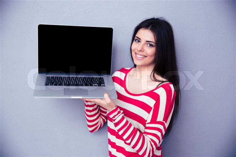 Smiling woman showing blank laptop display over gray background. Looking at camera, stock photo