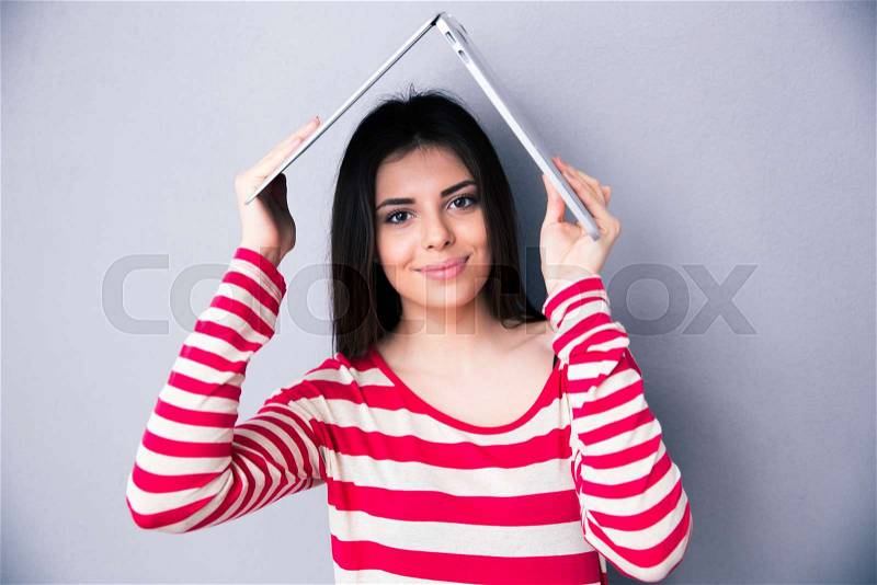 Pretty woman holding a laptop above her head like a roof over gray background. Looking at the camera, stock photo