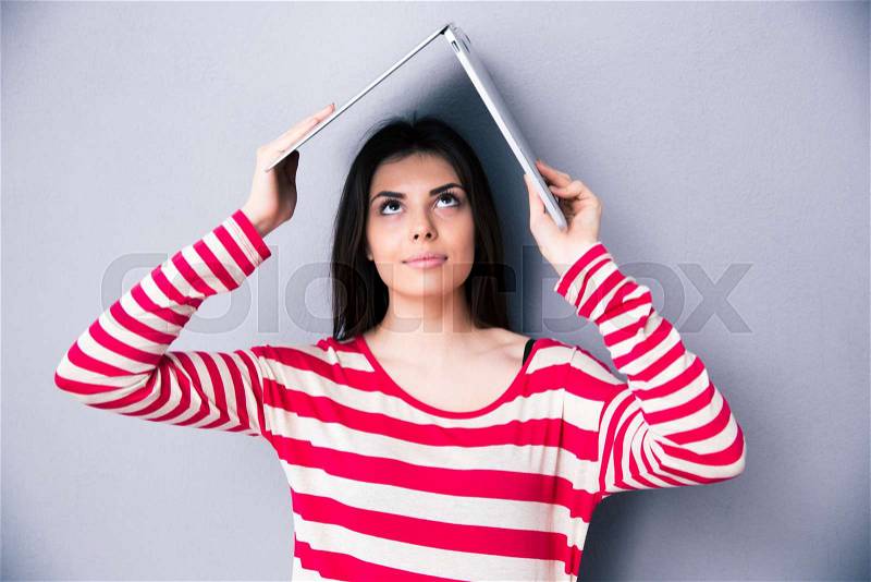 Beautiful woman holding laptop above her head like a roof over gray background. Looking up on laptop. , stock photo