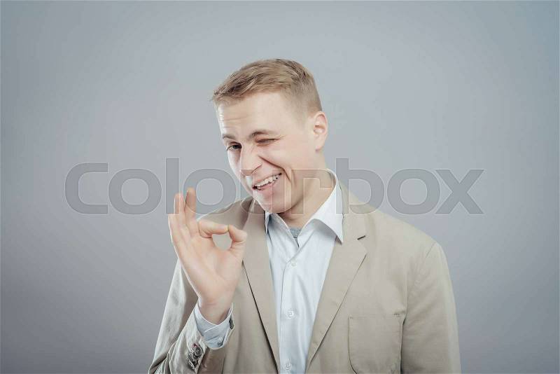 Gesturing OK sign. Cheerful young man in shirt gesturing OK sign while standing against grey background, stock photo