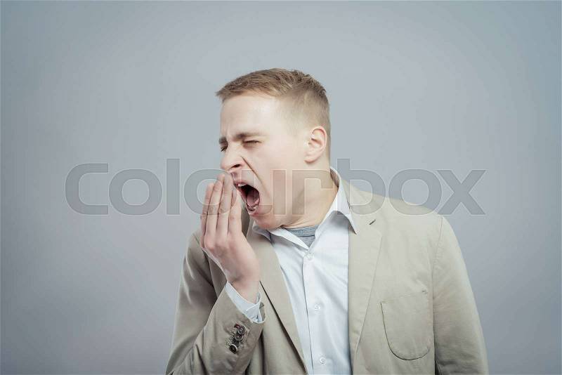 Closeup portrait young businessman funny guy placing hand on mouth yawning isolated grey wall background. Negative human emotion face expression feeling body language, stock photo