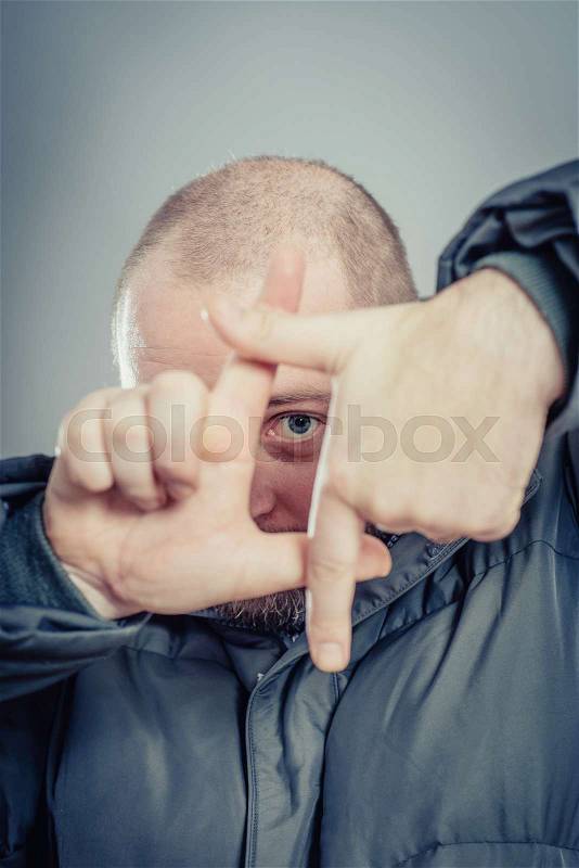Focusing at you. Playful young man in gesturing finger frame and smiling while standing against grey background, stock photo