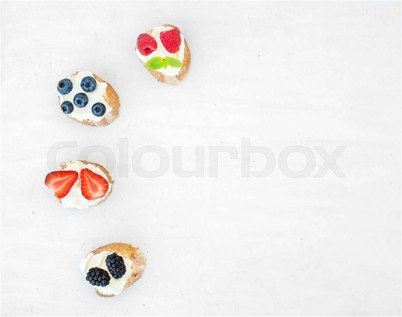 Sweet berry (strawberry, raspberry, blueberry and blackberry) and soft cheese sandwiches (bruschattas) over a white wooden background with a copy space. Top view, stock photo