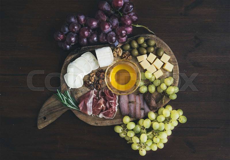 Wine appetizers set: meat and cheese selection, honey, grapes, walnuts and olives on a rustic wooden board over a dark wood background. Top view, stock photo