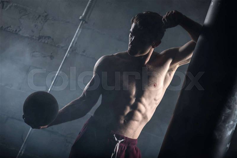 Young athlete with the ball in one hand stands near the punching bag. Snapshot in dark colors, stock photo