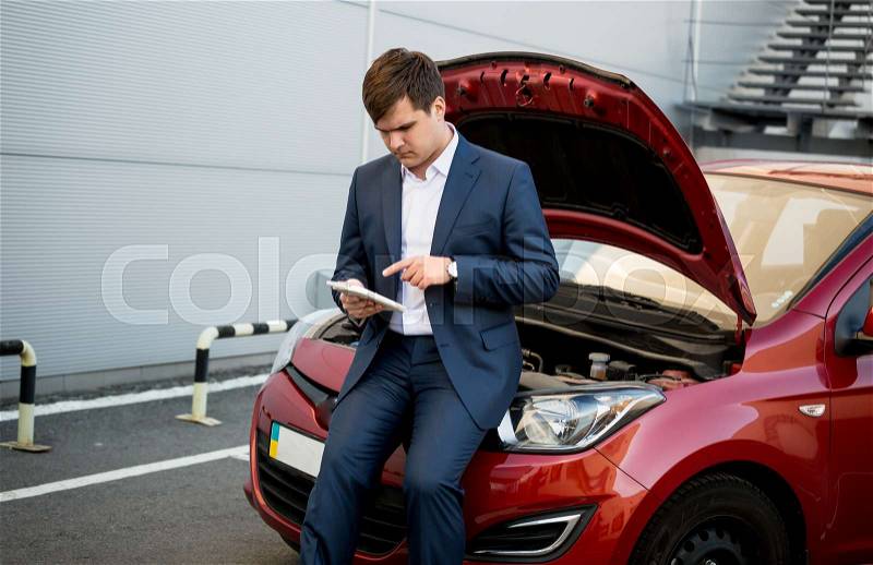 Young businessman sitting on hood of broken car and searching how to fix it on tablet, stock photo