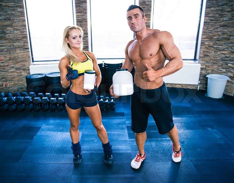 Muscular man and sporty woman holding container with sports nutrition and showing thumbs up in gym, stock photo