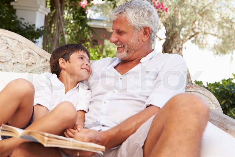 Grandfather And Grandson Sitting In Garden Reading Book, stock photo