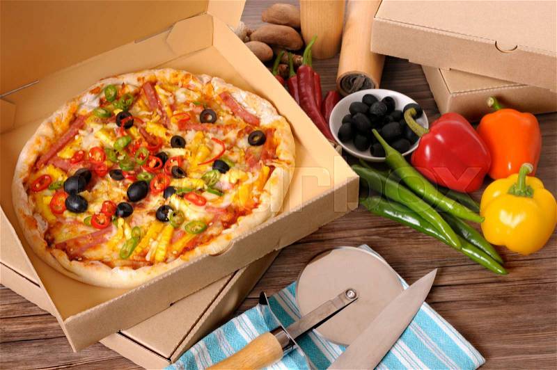 Freshly baked Pizza in a delivery box surrounded by various ingredients on a wood table or worktop, stock photo