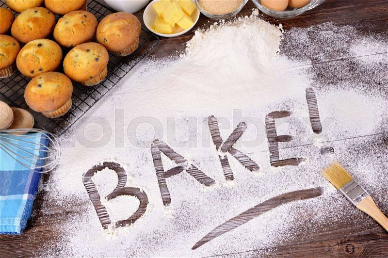 The word Bake written in flour on a dark wood table with freshly baked muffins and ingredients, stock photo