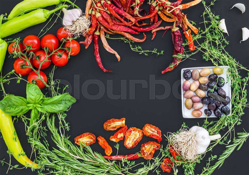 Mediterranean vegetable set consisting of garlic, chrry-tomatoes, dried tomatoes, fresh herbs, green and chili peppers and olives on a black surface with an empty space for you inscription in the center, stock photo