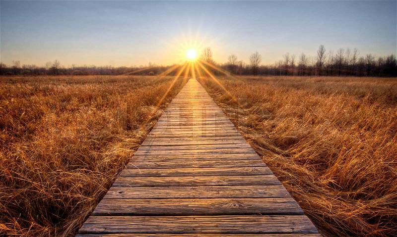 A beautiful sunset scene along a wood boardwalk with the boardwalk leading right into the setting sun. Find this boardwalk at Irwin Prairie State Nature Preserve in Northwest Ohio, stock photo