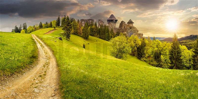 Composite mountain landscape. curve path to abandoned ruins of ancient fortress through green meadow on mountain hillside with forest in sunset light, stock photo