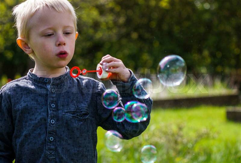 A Little boy blows soap bubbles in the garden on a summer day. He wears a blue shirt and is happy, stock photo