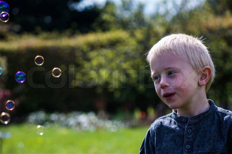 A Little boy blows soap bubbles in the garden on a summer day. He wears a blue shirt and is happy, stock photo