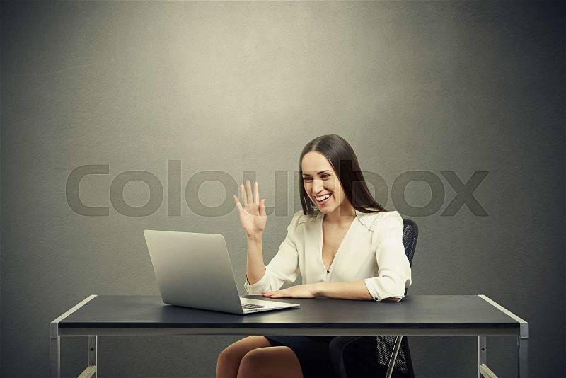 Smiley businesswoman have video chat, waving her hand and looking at laptop over dark background, stock photo