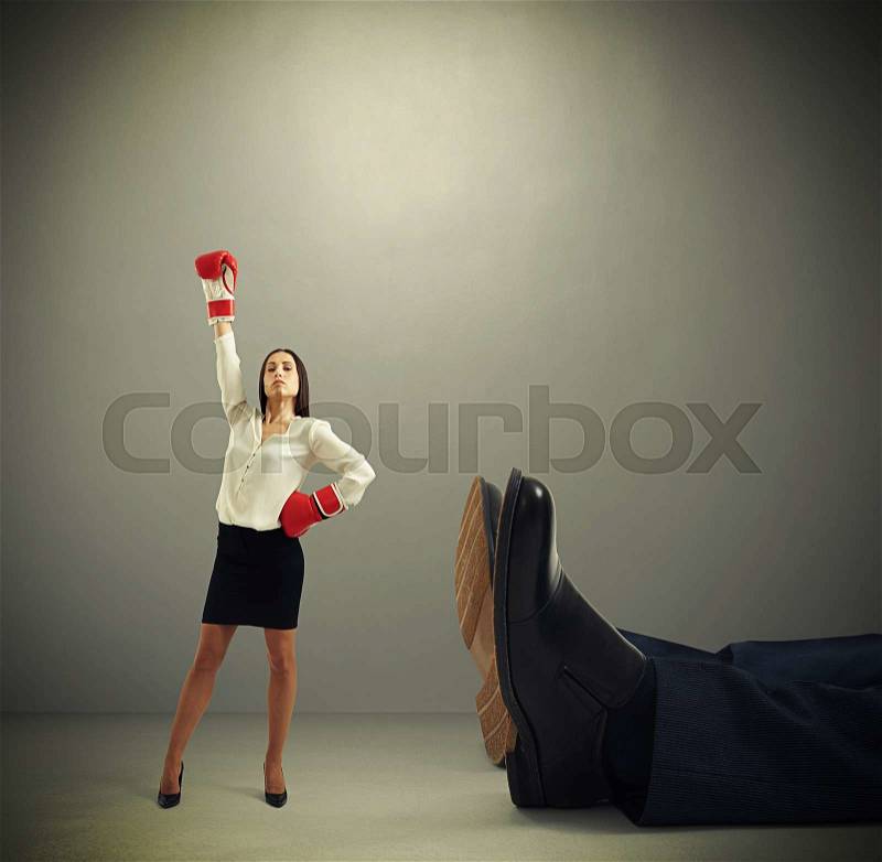 Winner businesswoman in red boxing gloves standing near big lying mans legs over dark grey background, stock photo