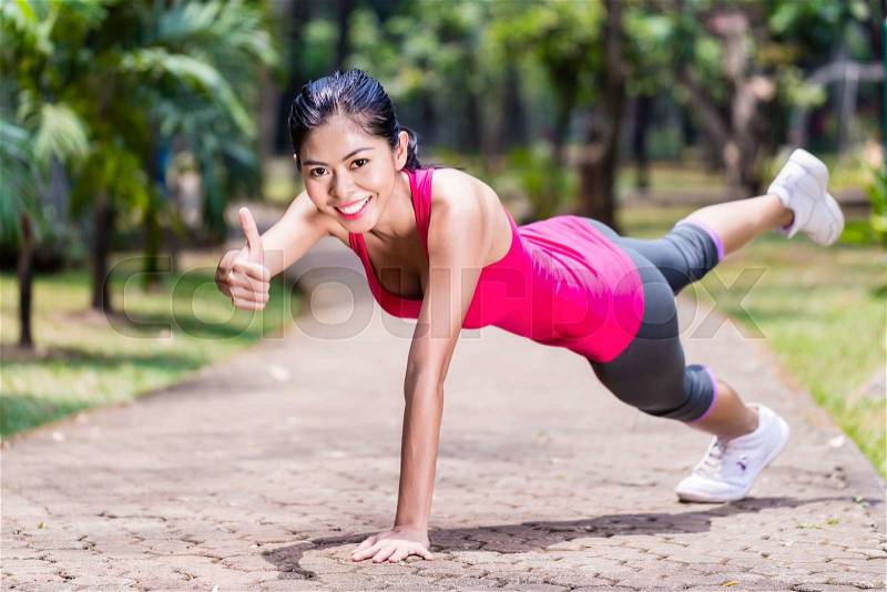 Proud and successful woman doing sport push-up in tropical Asian park, giving the thumbs up sign during her exercise, stock photo