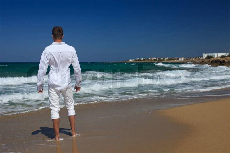 Barefoot man in a white clothing standing on the beach, view from the back, horizontal , stock photo