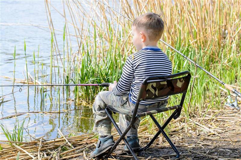 Rear View of Young Boy Sitting in Folding Chair at Edge of Pond and Fishing Patiently Amongst the Reeds, stock photo