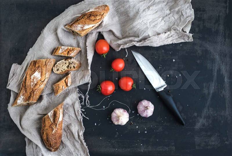 French baguette cut into pieces, cherry-tomatoes, garlic and kitchen knife over dark grunge background. Top view, stock photo