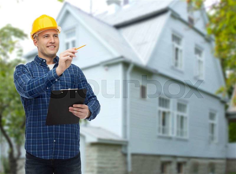 Repair, construction, building, people and maintenance concept - smiling male builder or manual worker in helmet with clipboard taking notes over living house background, stock photo