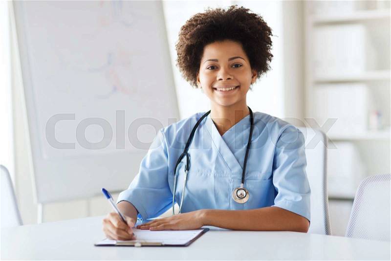 Medicine, people and healthcare concept - happy female african american doctor or nurse writing medical report to clipboard at hospital, stock photo