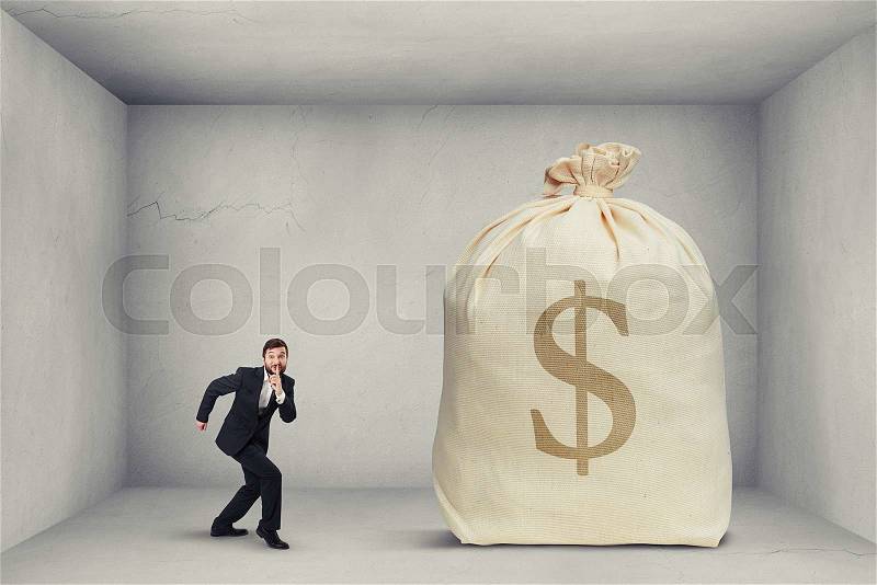 Businessman in formal wear showing silent sign and walking on tiptoe to the big bag of money in grey room , stock photo