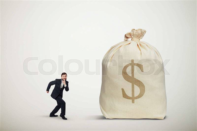 Businessman in formal wear showing silent sign and sneaking to the big bag of money over light grey background, stock photo