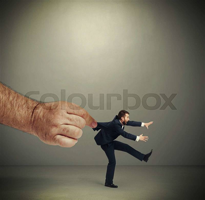 Big mans hand kepping emotional running businessman over dark grey background, stock photo
