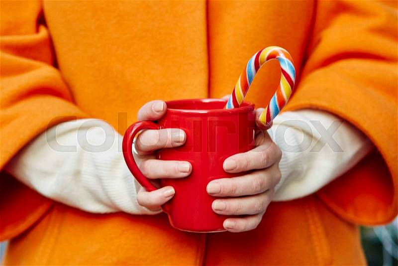 Woman hands in woolen mittens holding a cozy mug with hot cocoa, tea or coffee and a Christmas candy cane, stock photo