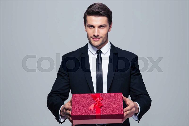 Handsome businessman holding gift box over gray background and looking at camera, stock photo