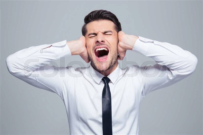 Businessman covering his ears and screaming over gray background, stock photo