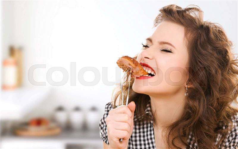 People, diet, culinary and food concept - hungry young woman eating meat on fork over kitchen background, stock photo